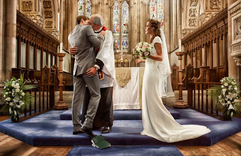 A man in a grey suit and a woman in a white wedding dress are kissing at the altar, with a priest in a white robe in between them.