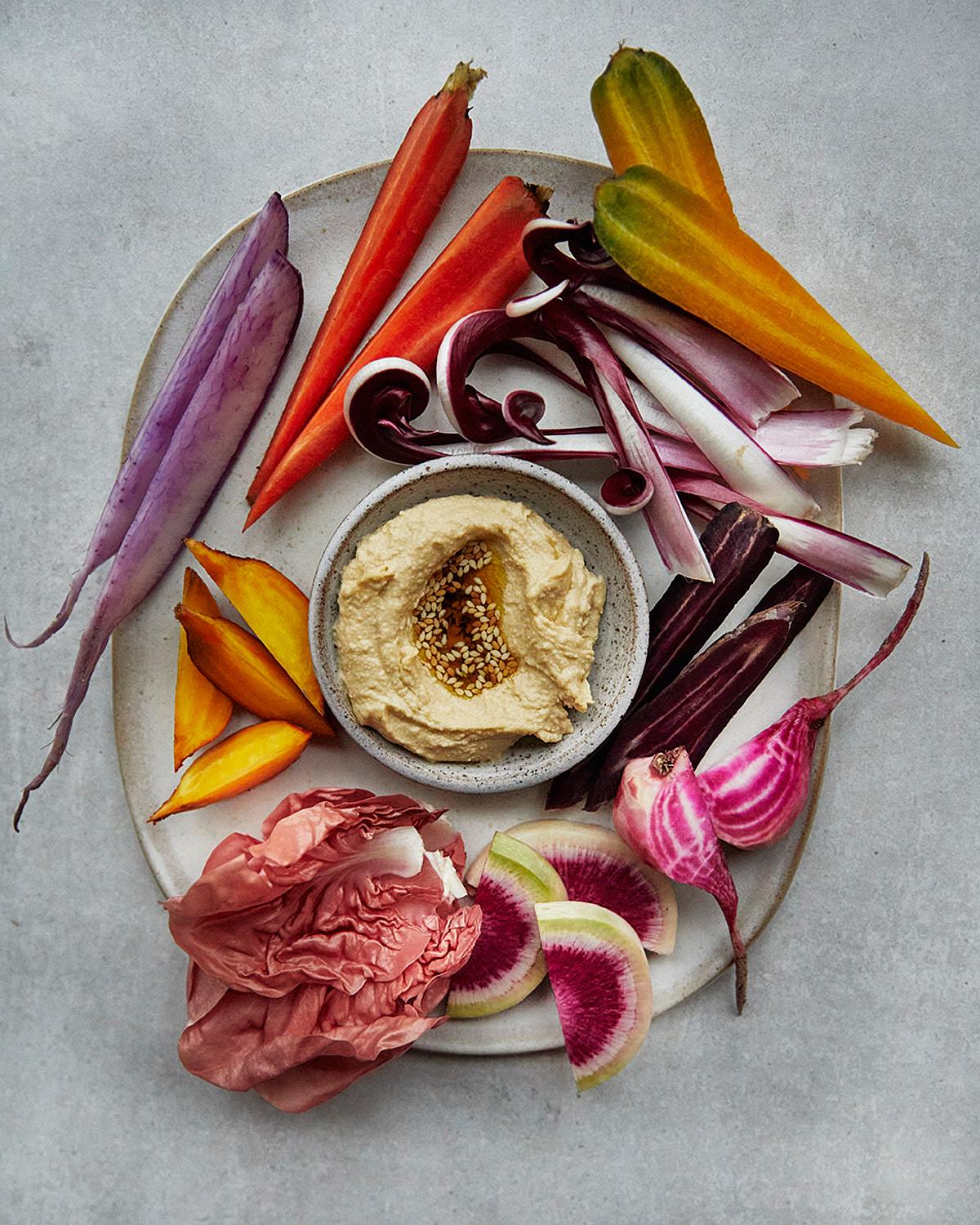 A selection of colorful, raw, and fresh vegetables and a bowl of hummus on a white plate.