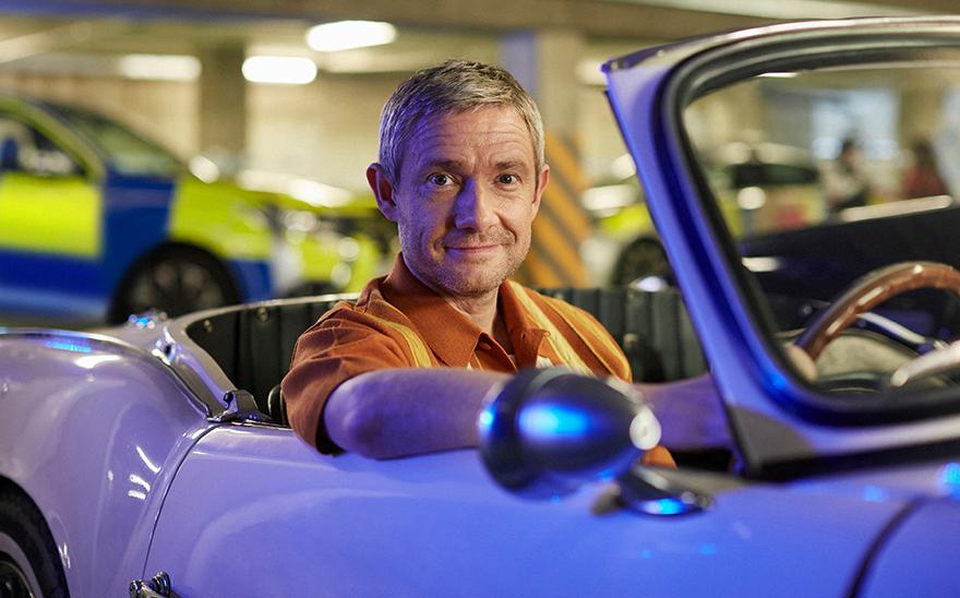 A man in an orange shirt is sitting in a white sports car in a garage or similar setting, with a police car in the background.