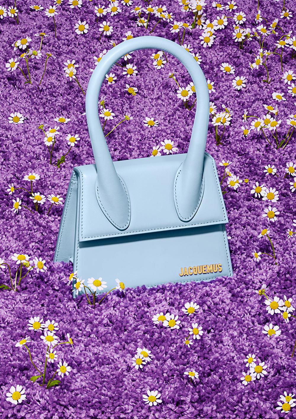 A small, light blue handbag with a handle, surrounded by a bed of purple flowers and white daisies.