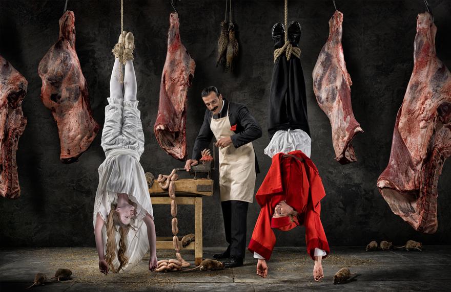 A man in a black suit and white apron stands in a meat market surrounded by hanging meat while two women do handstands on the floor.