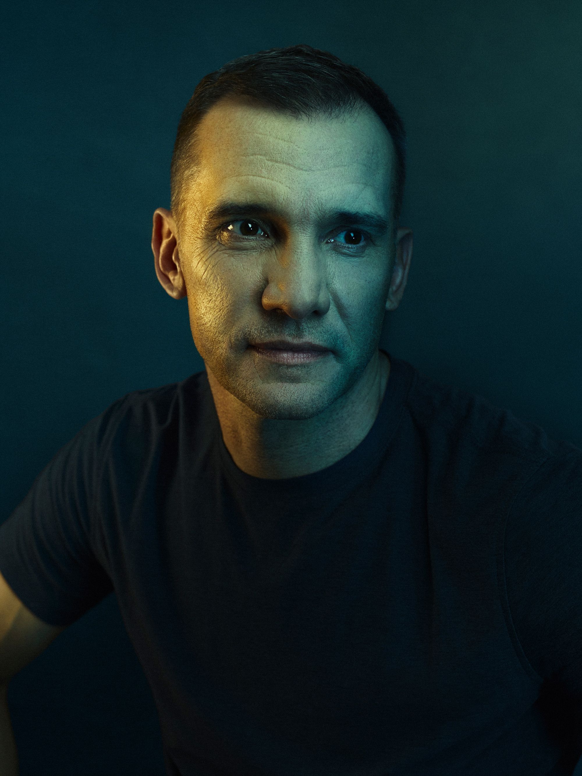 A man with a serious expression, wearing a black t-shirt, in a dark, moody environment.