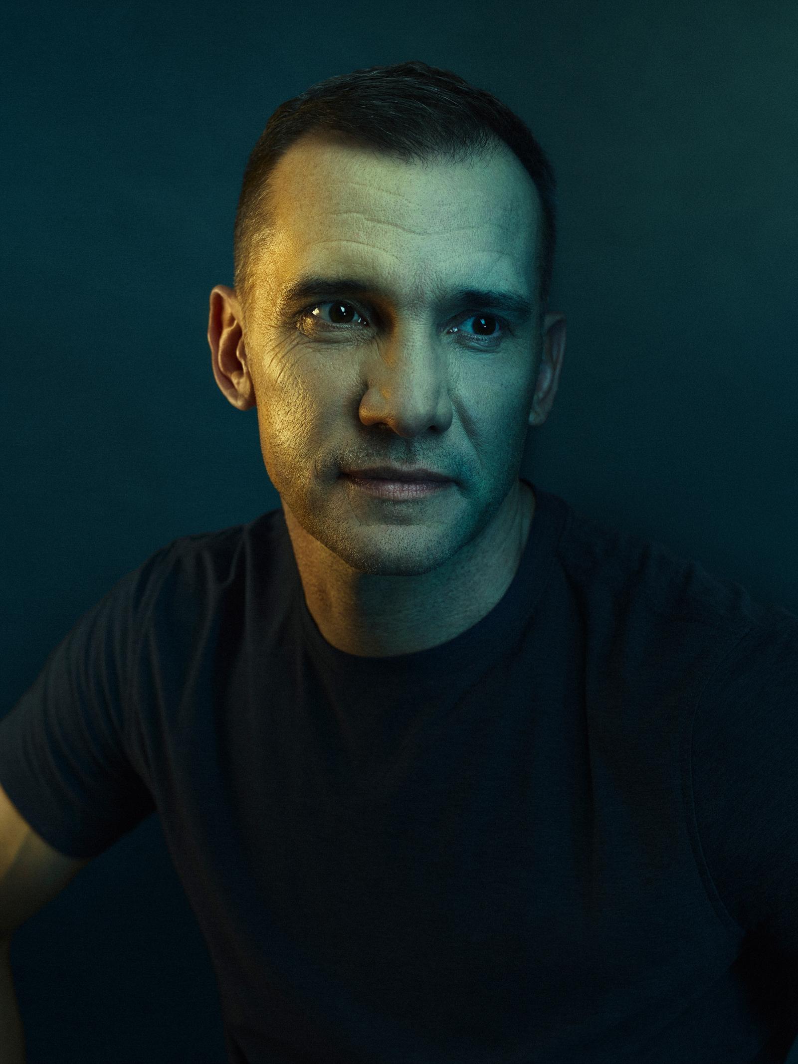 A man with a serious expression, wearing a black t-shirt, in a dark, moody environment.