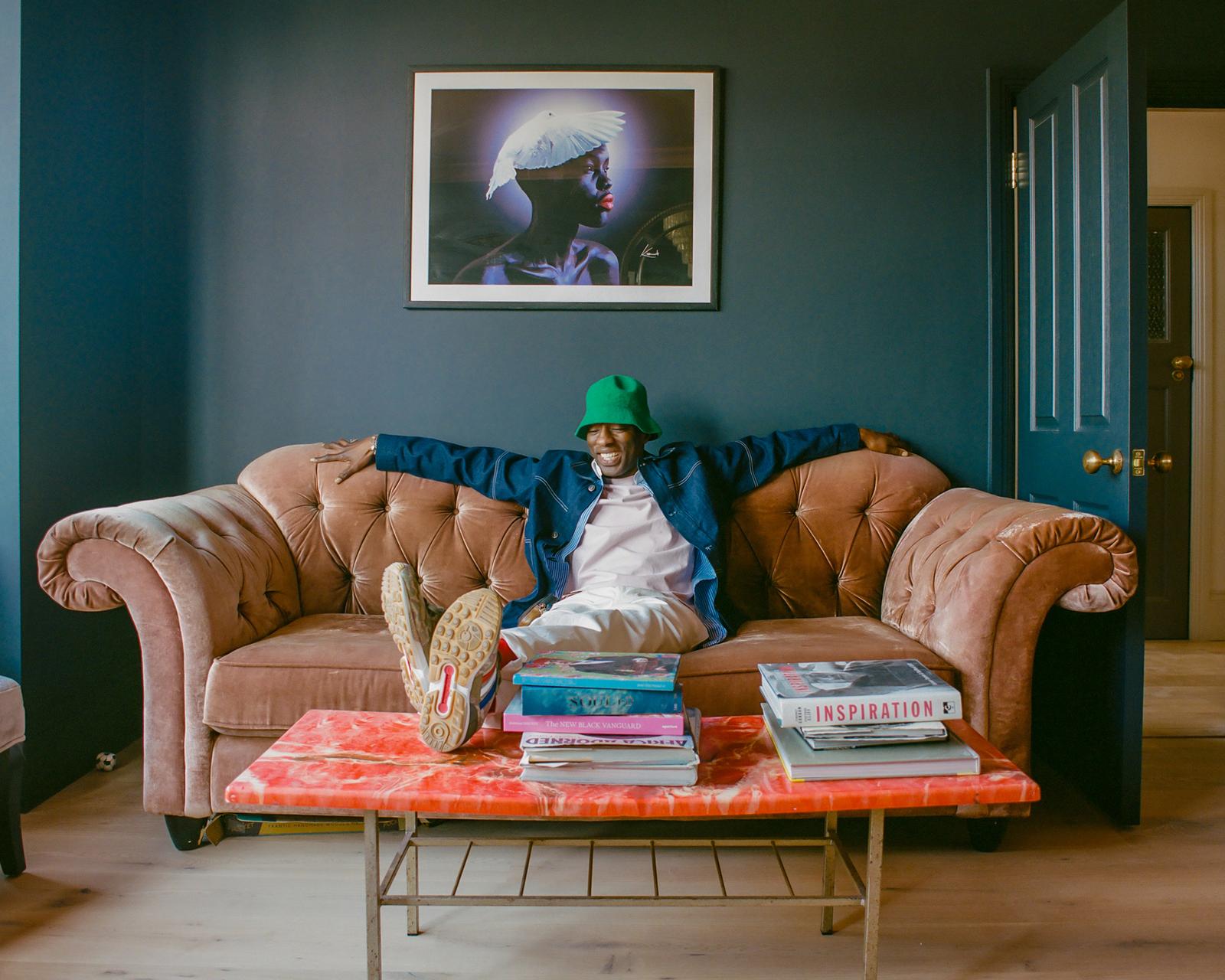 A man in a green hat and denim jacket is sitting on a couch, surrounded by books and a coffee table with a red marble top.