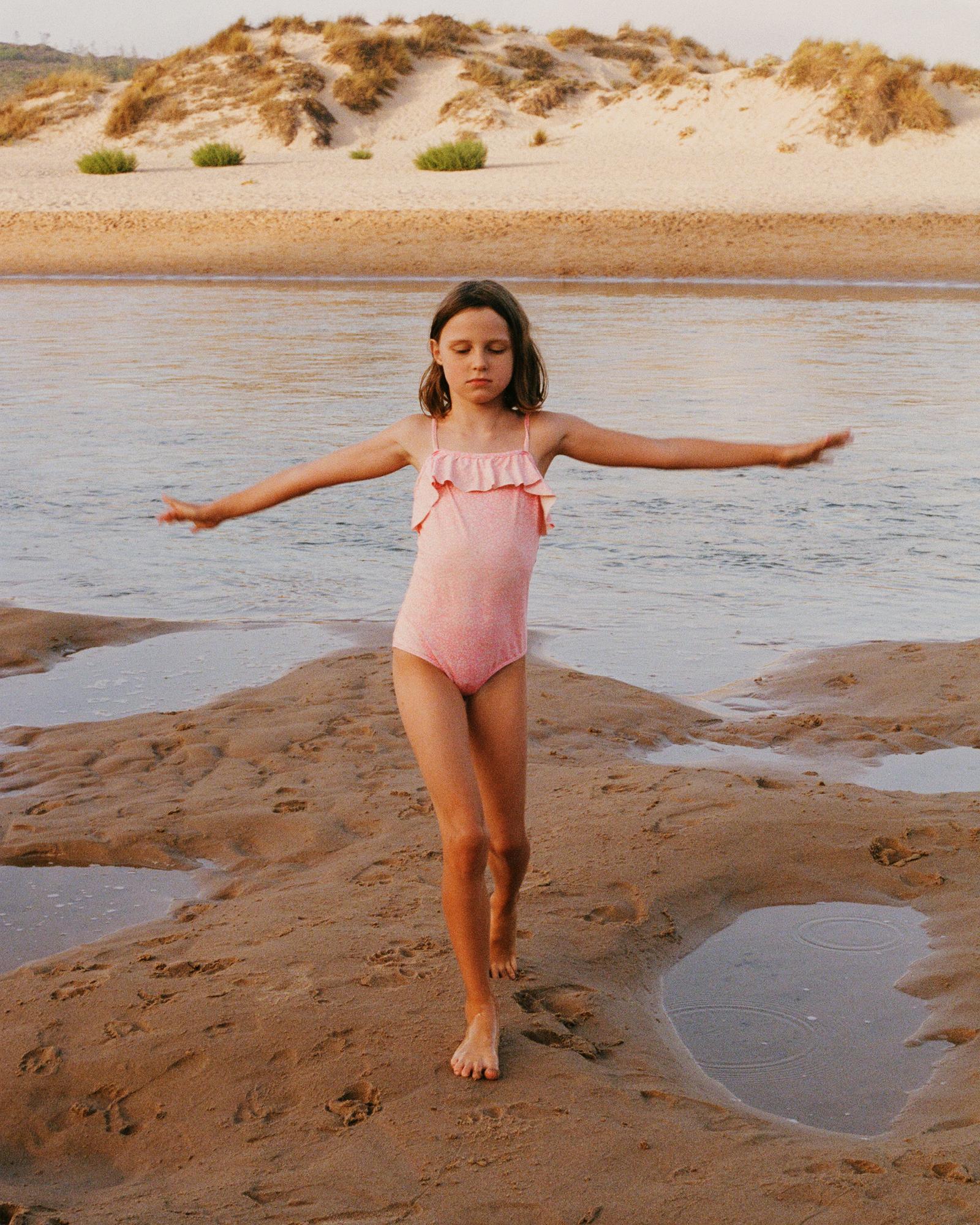 A young girl in a pink swimsuit is running on a sandy beach, with her arms outstretched and a joyful expression on her face.