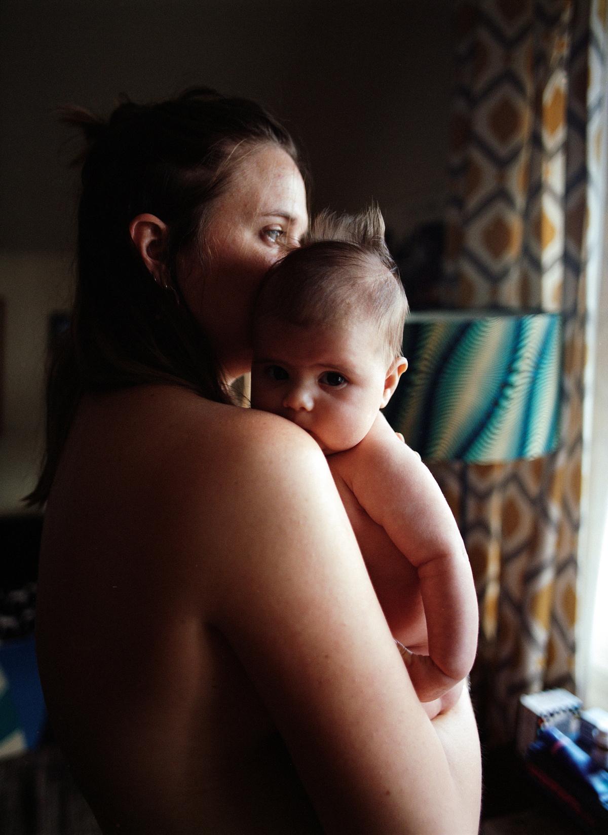 A woman with her back to the camera holds a baby in a dimly lit room.