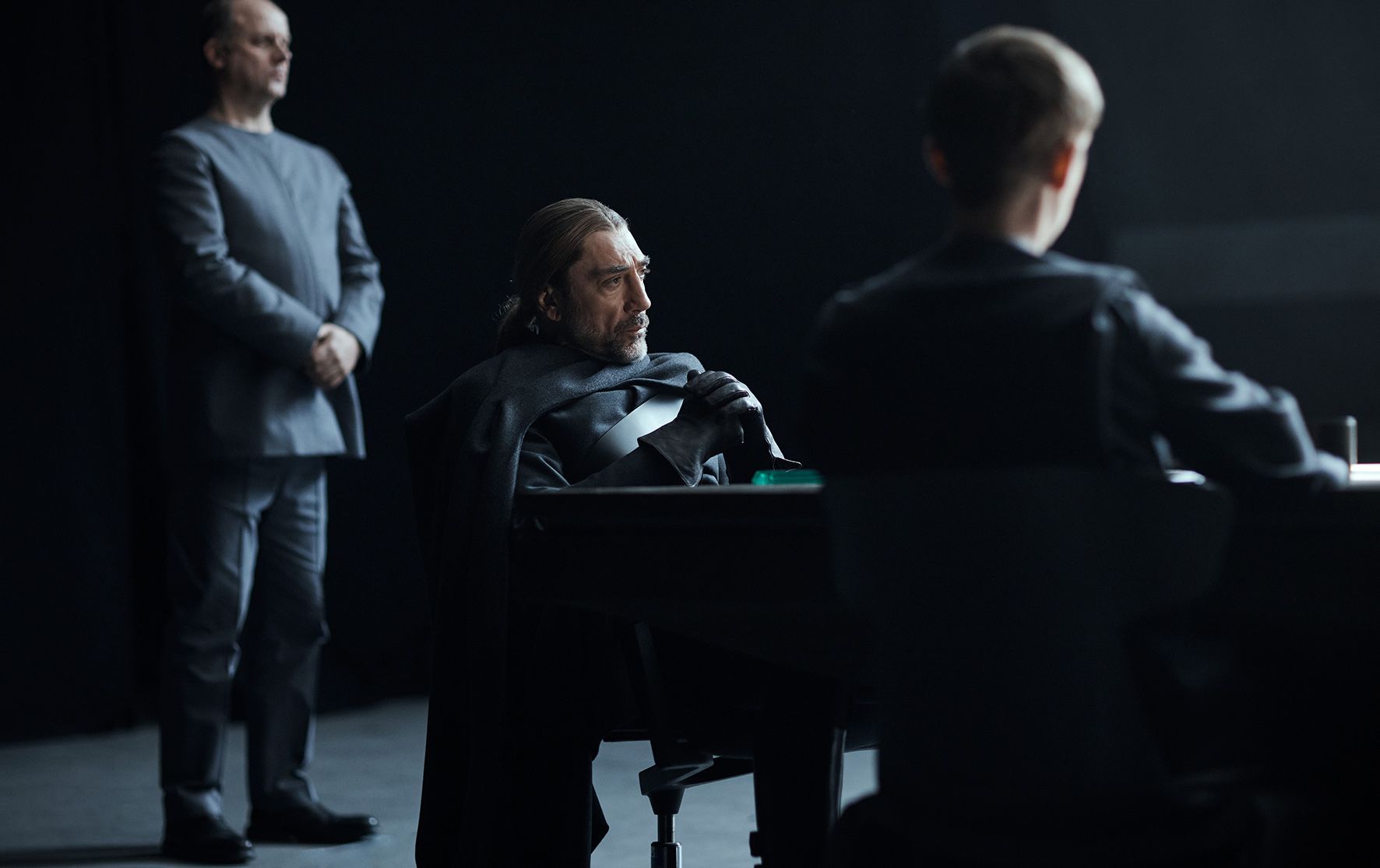 Javier Bardem sits at a table, looking at a screen, with two men in the background in a dark, modern room.