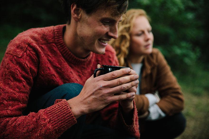 A young man in a red sweater and a woman in a brown jacket are sitting on the ground, holding a cup and looking at something in the distance.