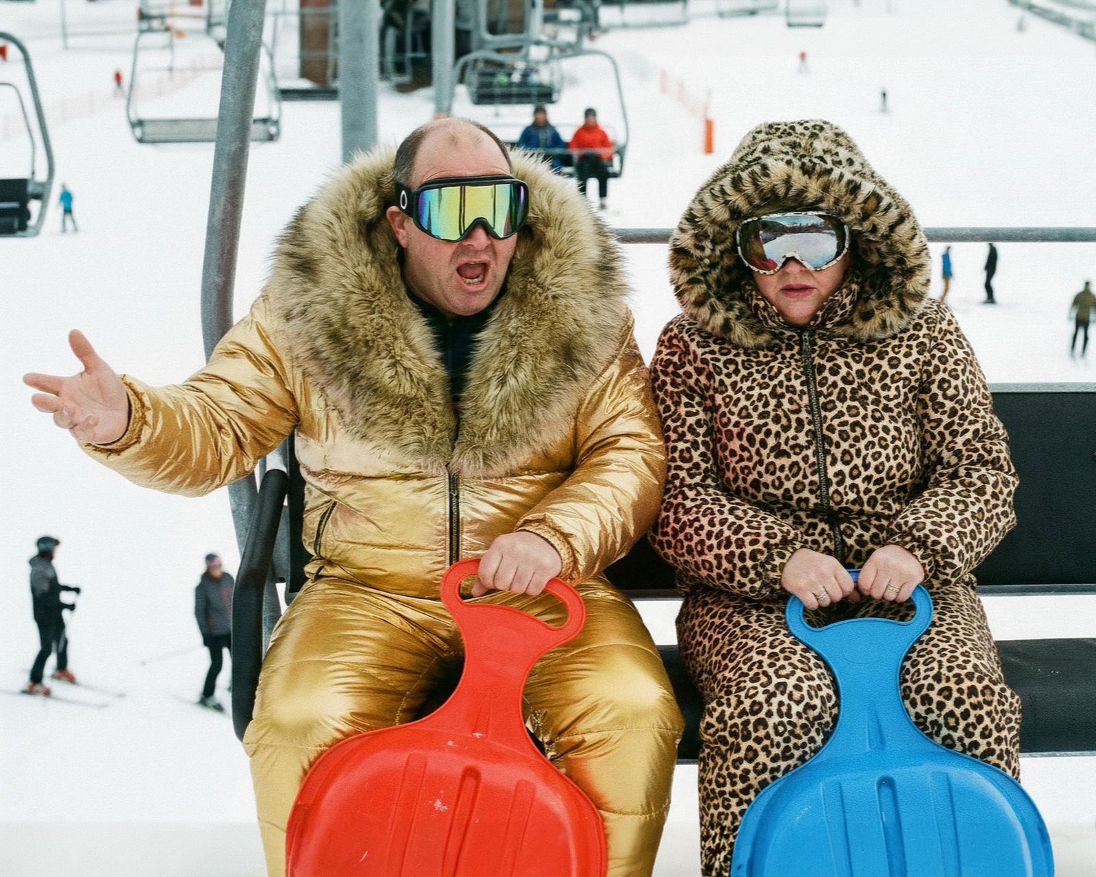 A man and a woman, both wearing winter clothing, sitting on a bench in a snowy landscape, with a ski lift in the background.