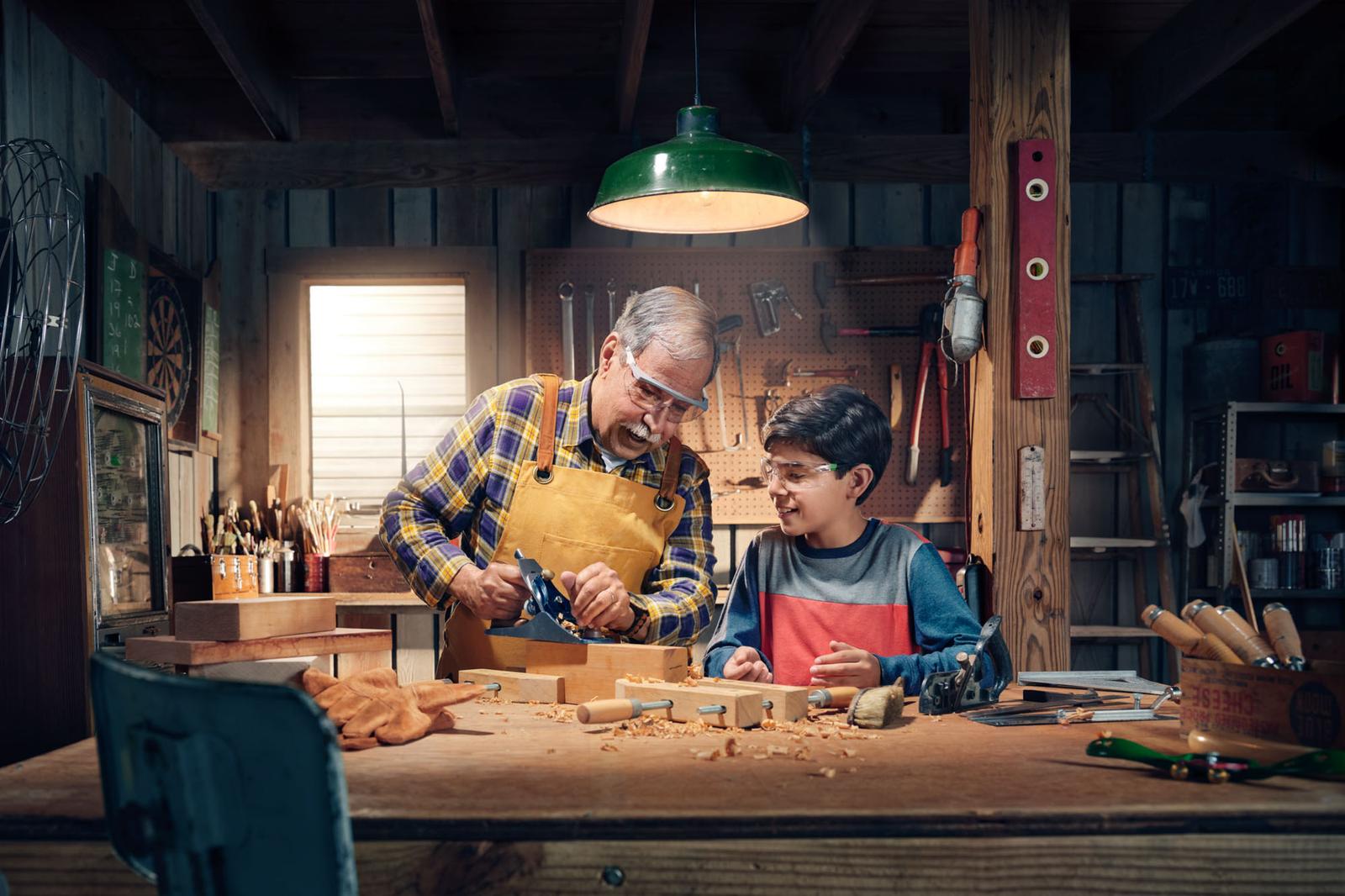 An older man with a mustache and a young boy in a workshop, both wearing safety glasses and working with a hand plane on a piece of wood.
