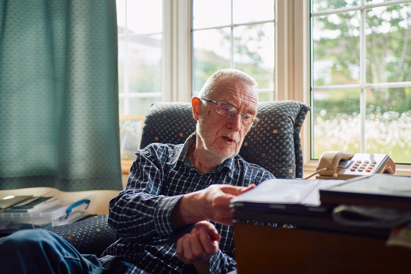 An older man sits in a chair wearing a blue and white plaid shirt and glasses.