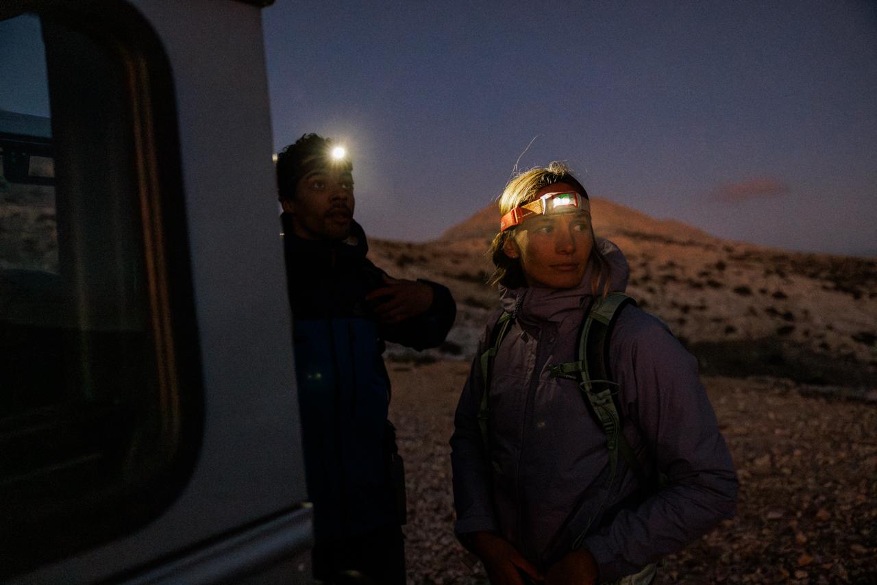 Two people, one male and one female, standing in a desert-like landscape at night, both wearing headlamps and backpacks, with a vehicle in the background.