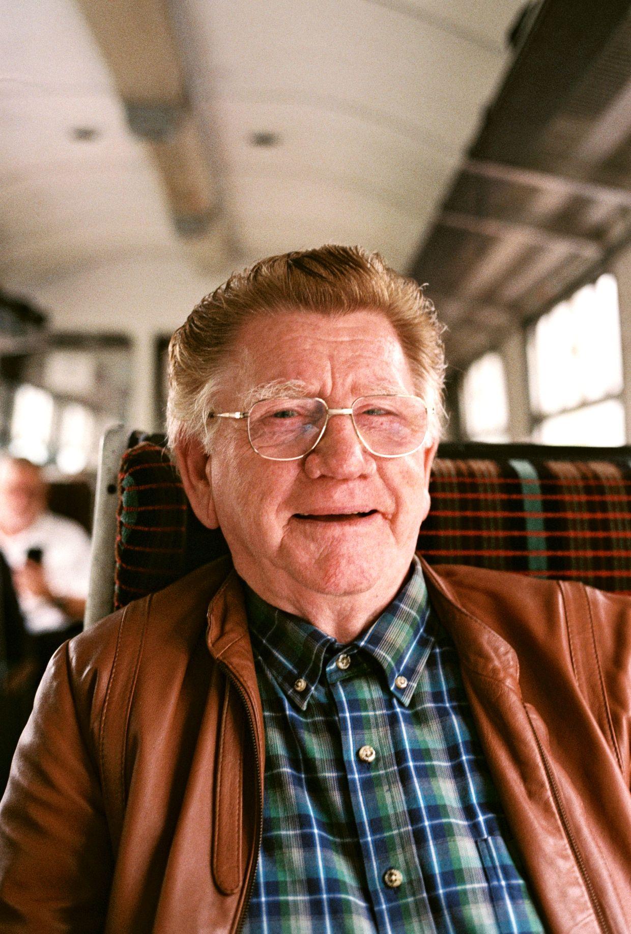 An older man with a warm, gentle smile sits in a train or bus, wearing a brown leather jacket over a blue and green plaid shirt, with a blurred view of a city behind him.