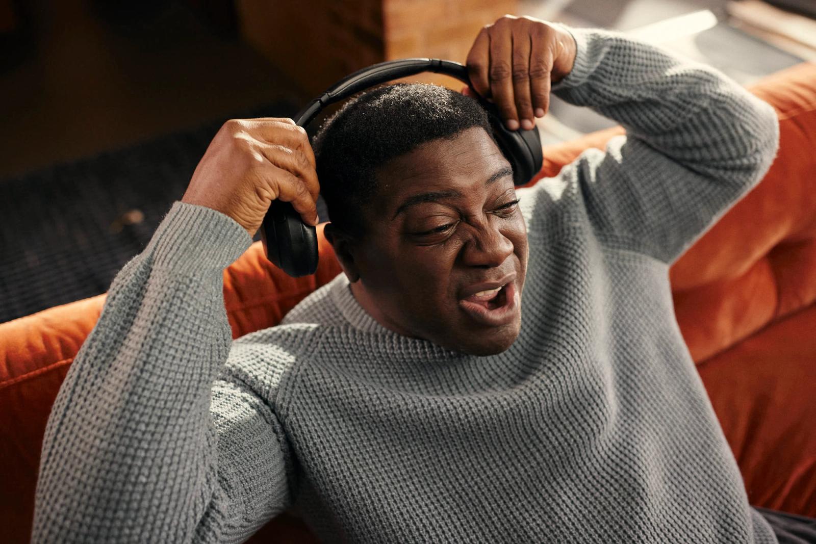 A man wearing a grey sweater sits on a red couch, holding his headphones to his head as he listens to music. He has a relaxed, happy expression on his face, with his eyes closed.