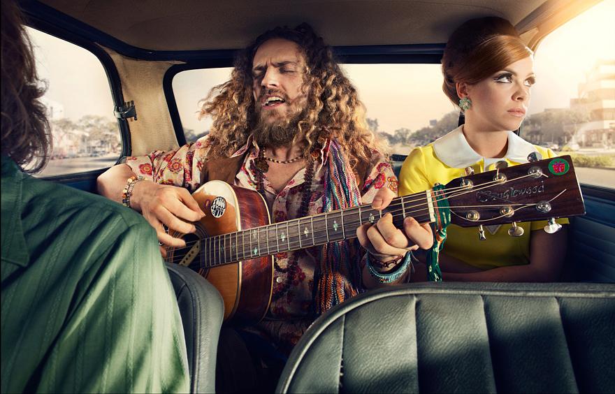 A man with long, curly hair and a woman with a red hair bun are in a car, the man is playing a guitar and the woman is looking out the window.