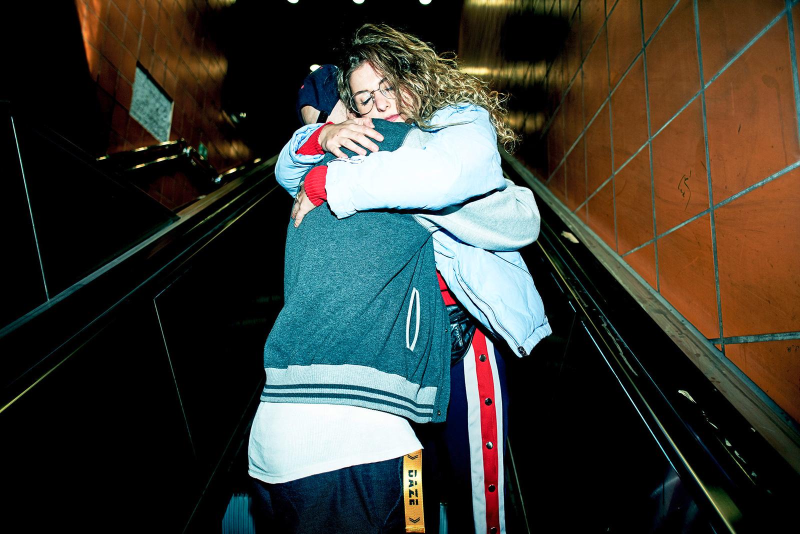Two people embracing on an escalator in a dimly lit, underground setting, both wearing casual, streetwear-inspired clothing.
