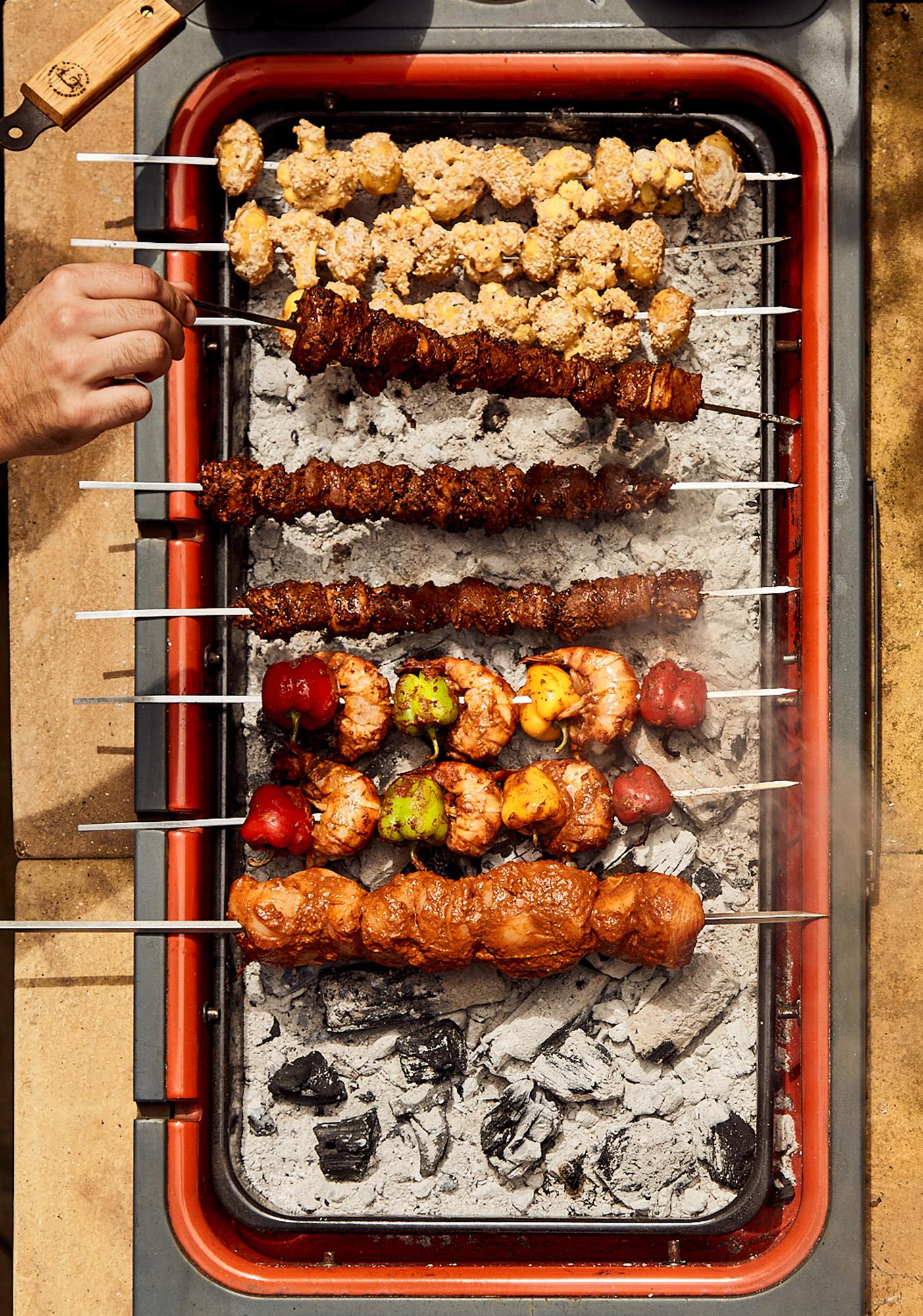 A person's hand is holding a metal skewer with a variety of grilled meats, including beef, chicken, and vegetables, over a charcoal grill.