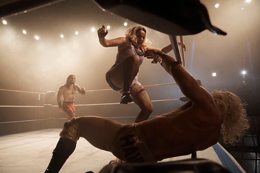A woman in a wrestling ring, wearing a pink tank top and black fishnet stockings, is being thrown by a wrestler in a gold and black wrestling costume.