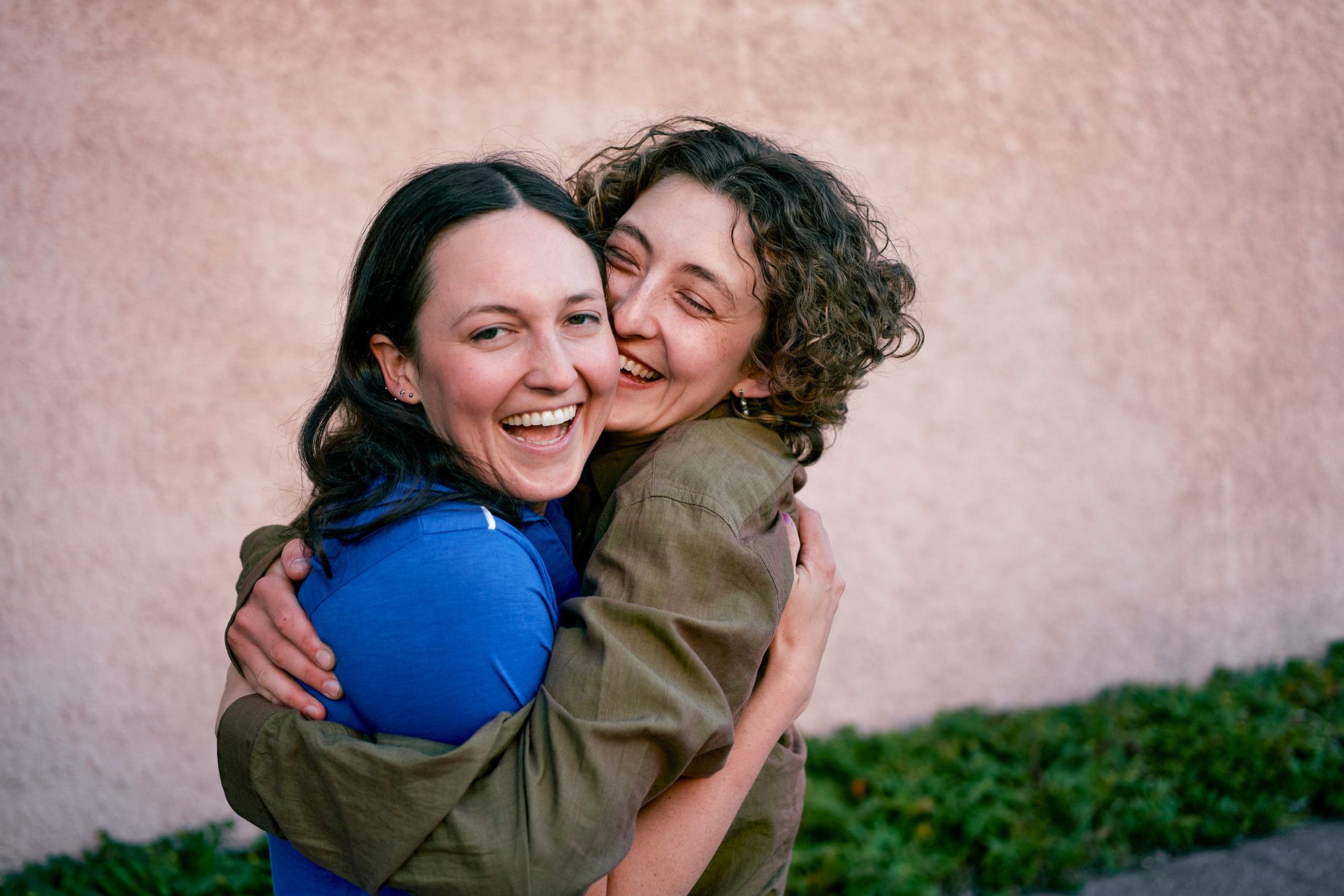 Two women embracing, smiling and laughing together, with one woman wearing a blue shirt and the other wearing a green shirt.