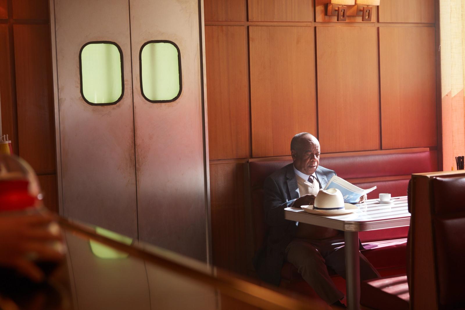 A man in a suit sits in a diner, reading a newspaper.