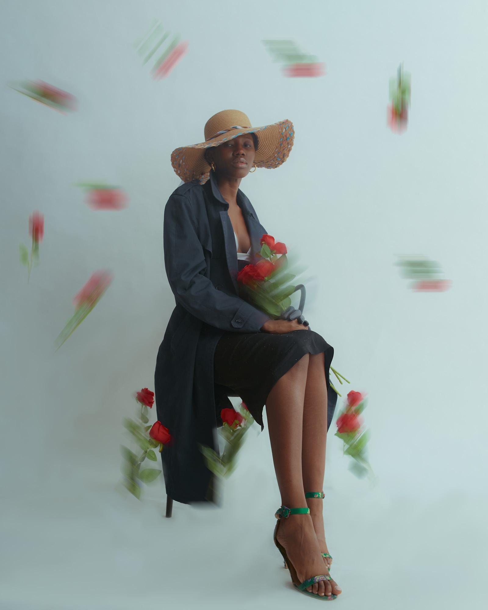A woman wearing a large, wide-brimmed straw hat, a black trench coat, and a white top, sitting on a chair, surrounded by falling red roses.