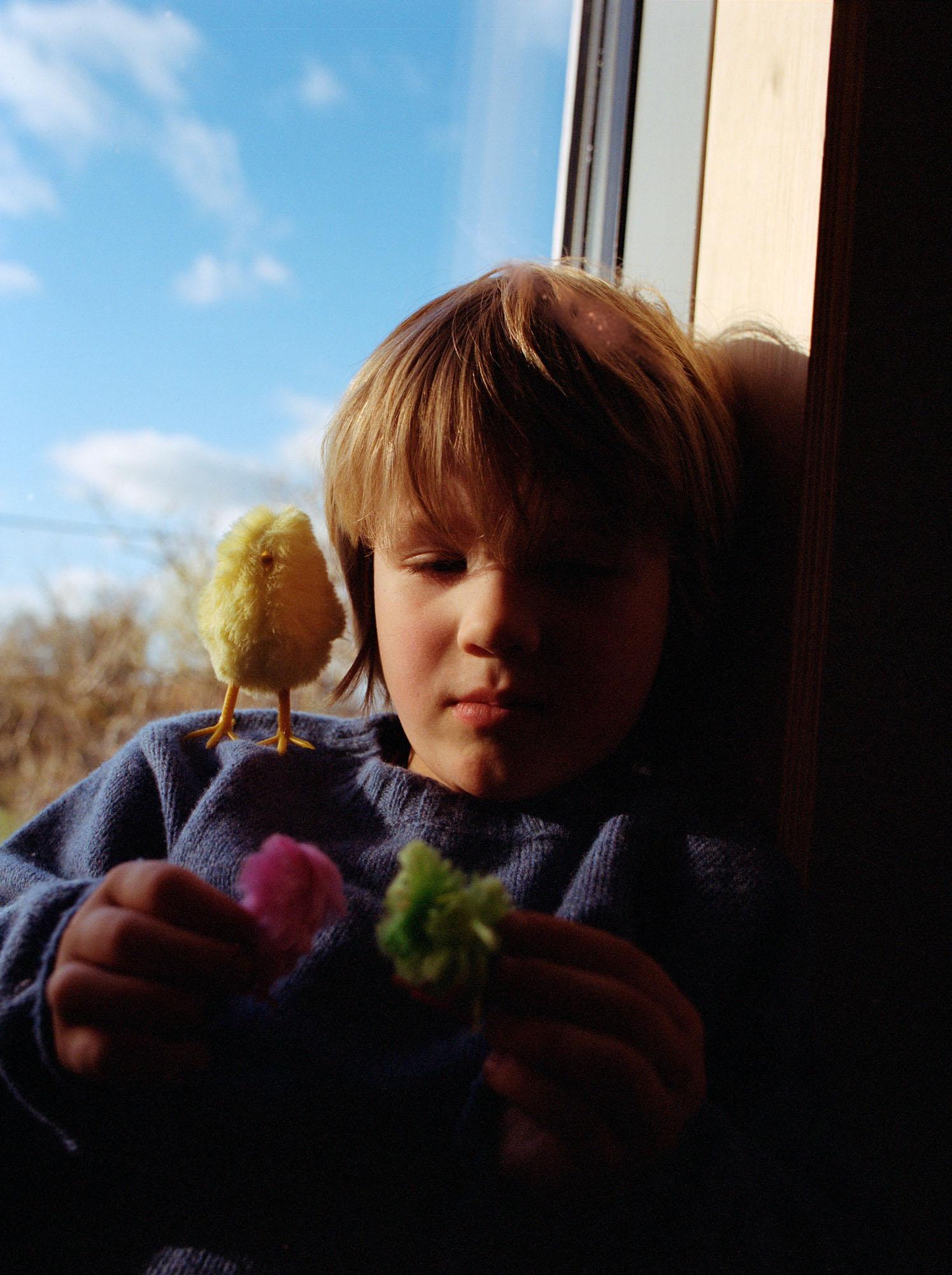 A young girl holds a yellow toy chick while wearing a blue sweater and a blue glove near a window with a blue sky and clouds.
