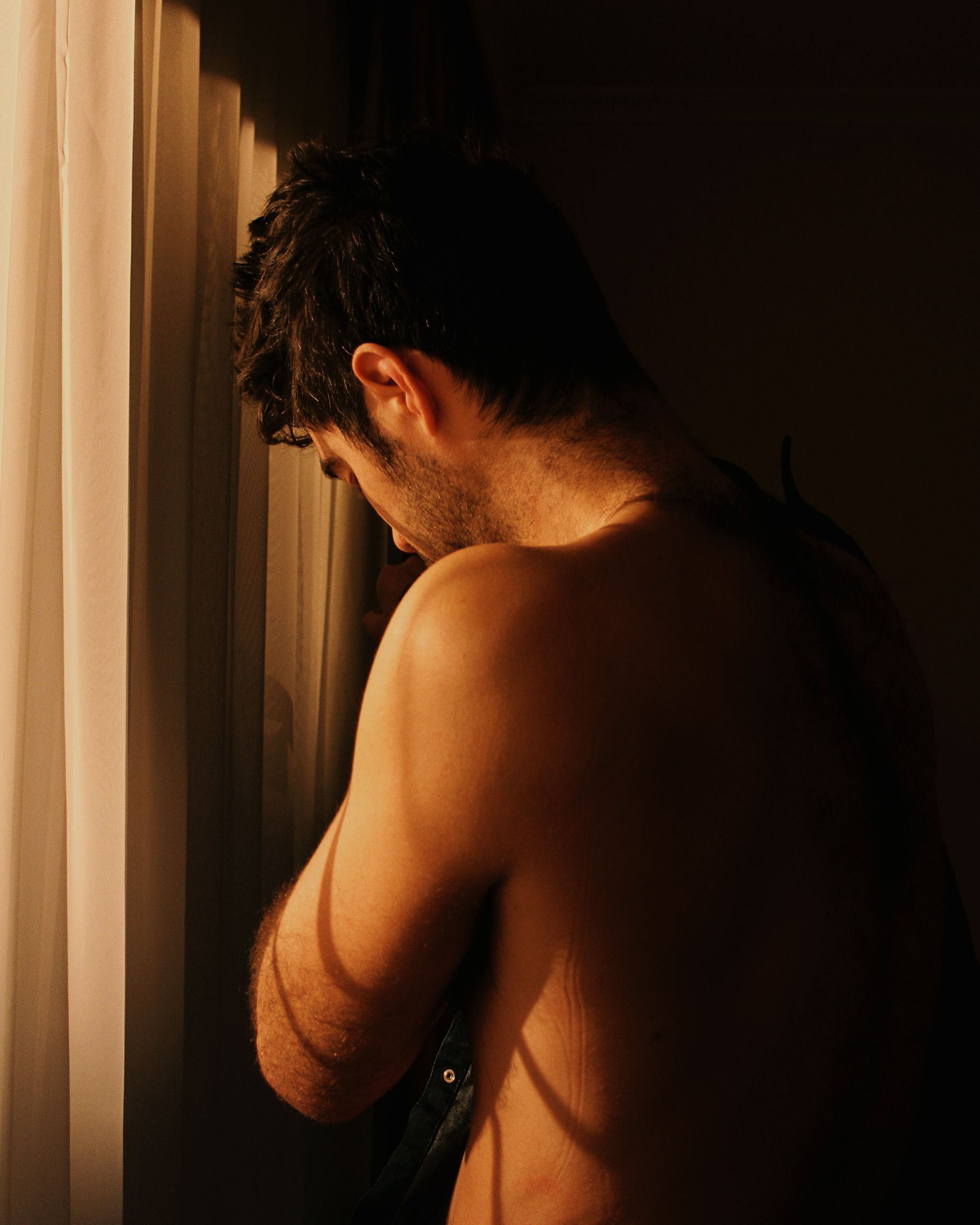 A shirtless man with his back to the camera, standing in front of a window with curtains, looking out at something in the distance.