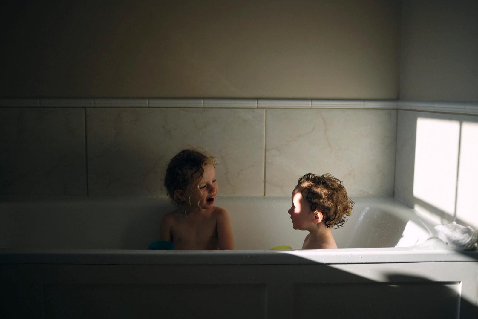 Two young children in a bathtub, laughing and playing with soap bubbles. The mood is playful and joyful.