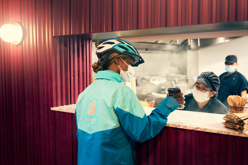 A food delivery person in a blue and green jacket, wearing a helmet and a mask, holding a phone and standing in front of a kitchen counter with a chef and a cook in the background.