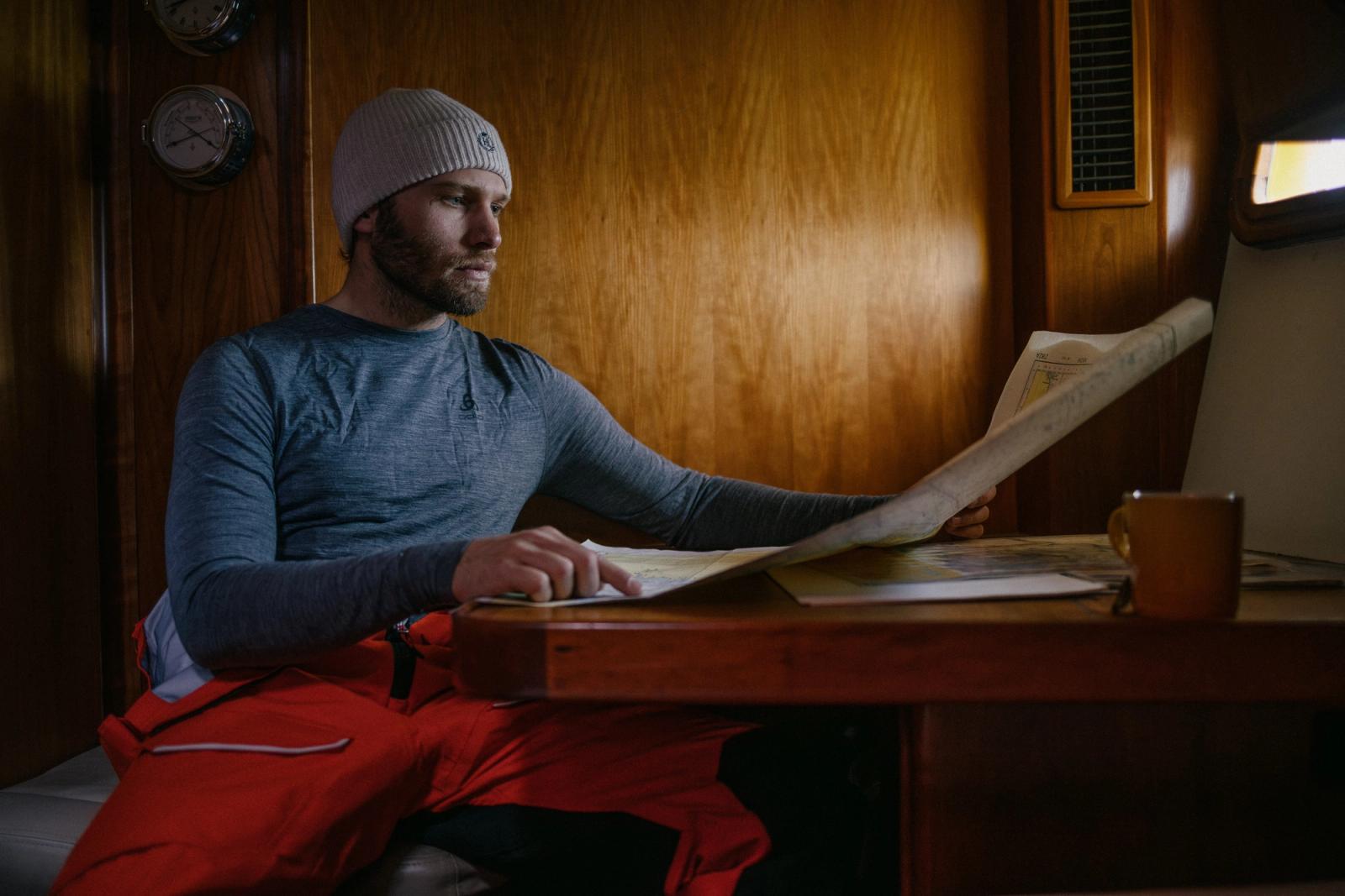 A man in a grey beanie, blue long-sleeved shirt, and red snow pants sits at a wooden table, studying a map.