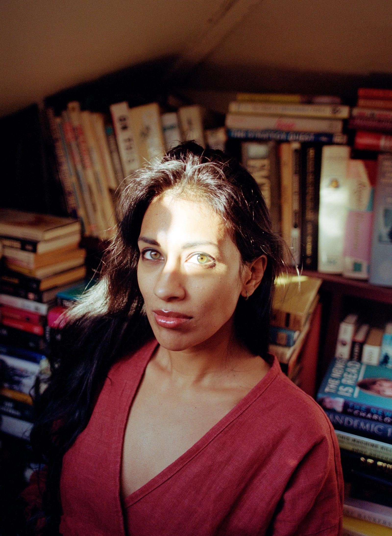 A woman with long dark hair and a red top in front of a bookshelf.