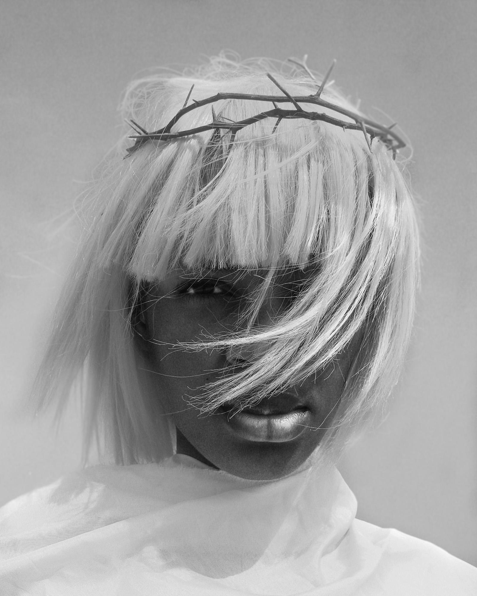 A person with a crown of thorns, likely a reference to the biblical story of Jesus Christ, wearing a white robe and a crown of thorns, with long hair and a serious expression.