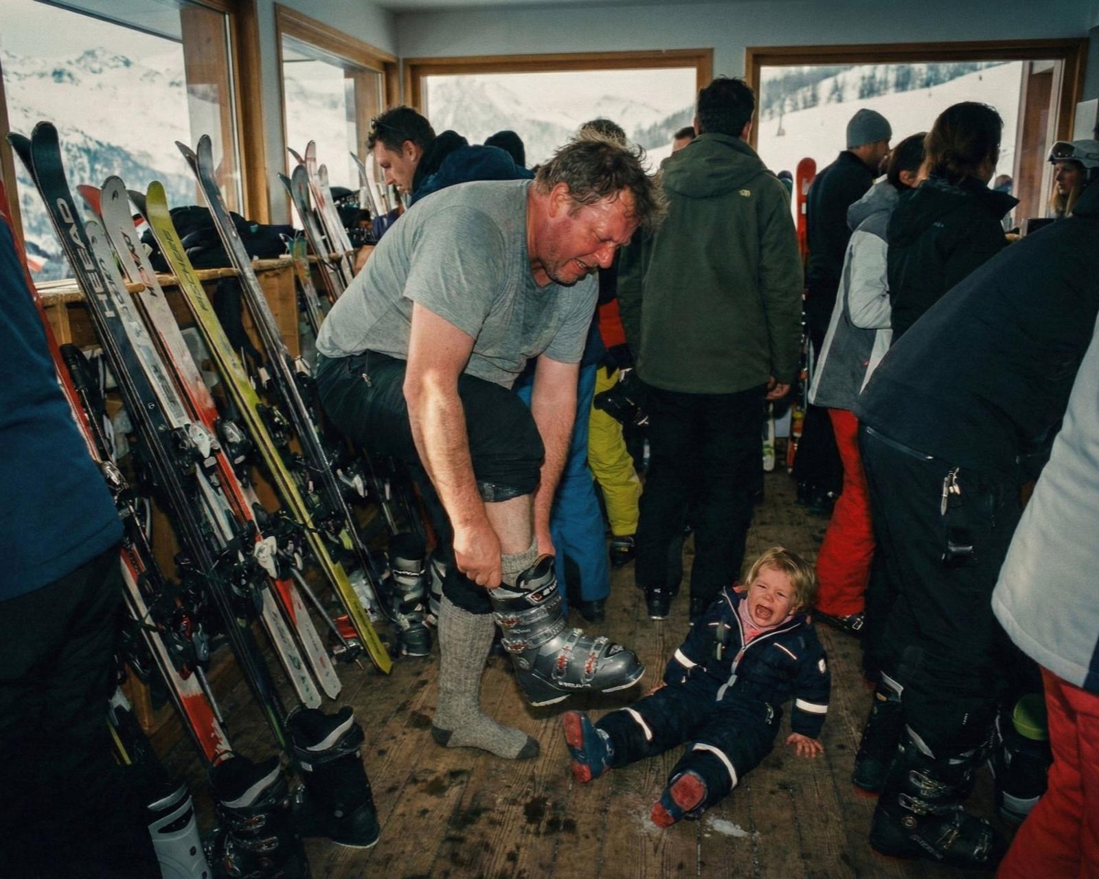 A man in a grey t-shirt and black shorts is helping a young child, who is wearing a black ski suit, to put on a pair of ski boots.