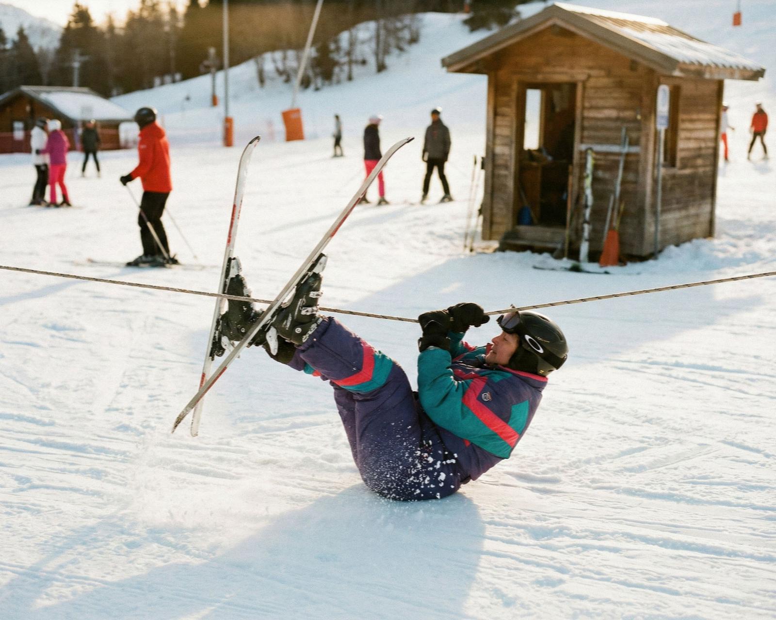 A person in a colorful ski suit is lying on their back in the snow, holding onto a ski lift cable with both hands.