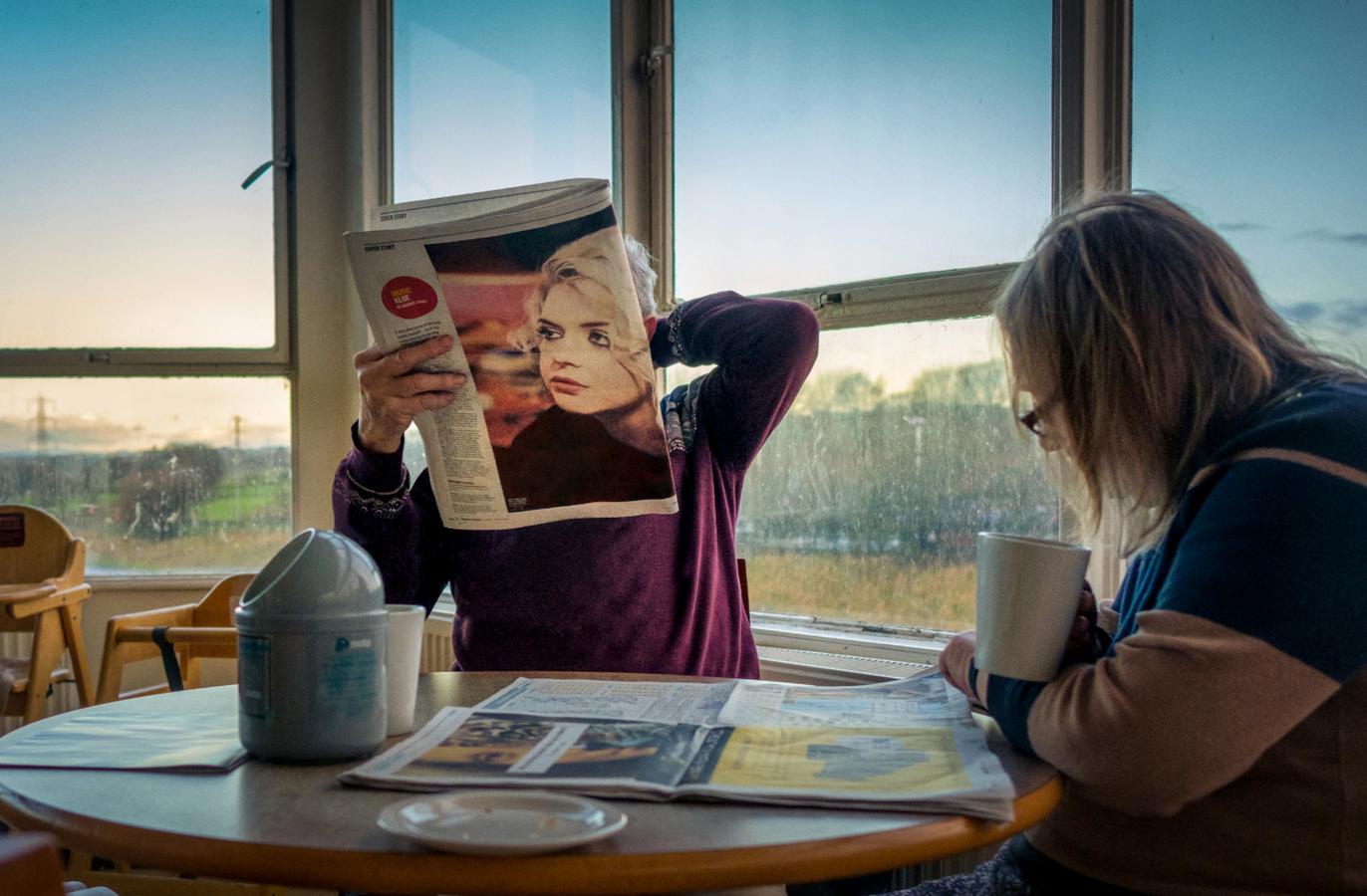 Two women sit at a table by a window; one reads a newspaper while the other holds a cup of coffee.