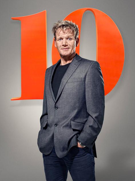 Gordon Ramsay stands in front of a large '24' logo wearing a grey suit jacket, black t-shirt, and a watch.