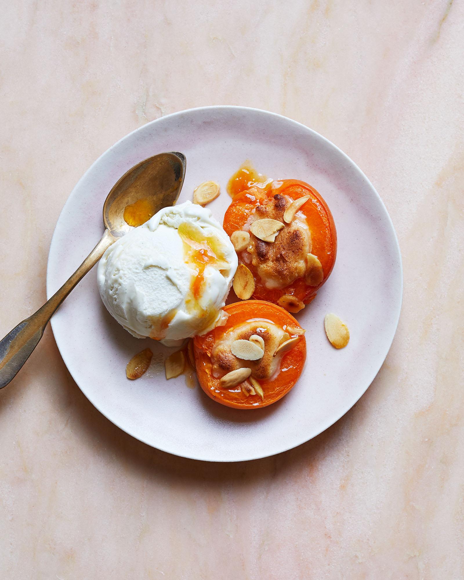 A dessert plate with a scoop of ice cream, two slices of baked peaches, and a drizzle of honey, served on a white plate with a gold spoon.