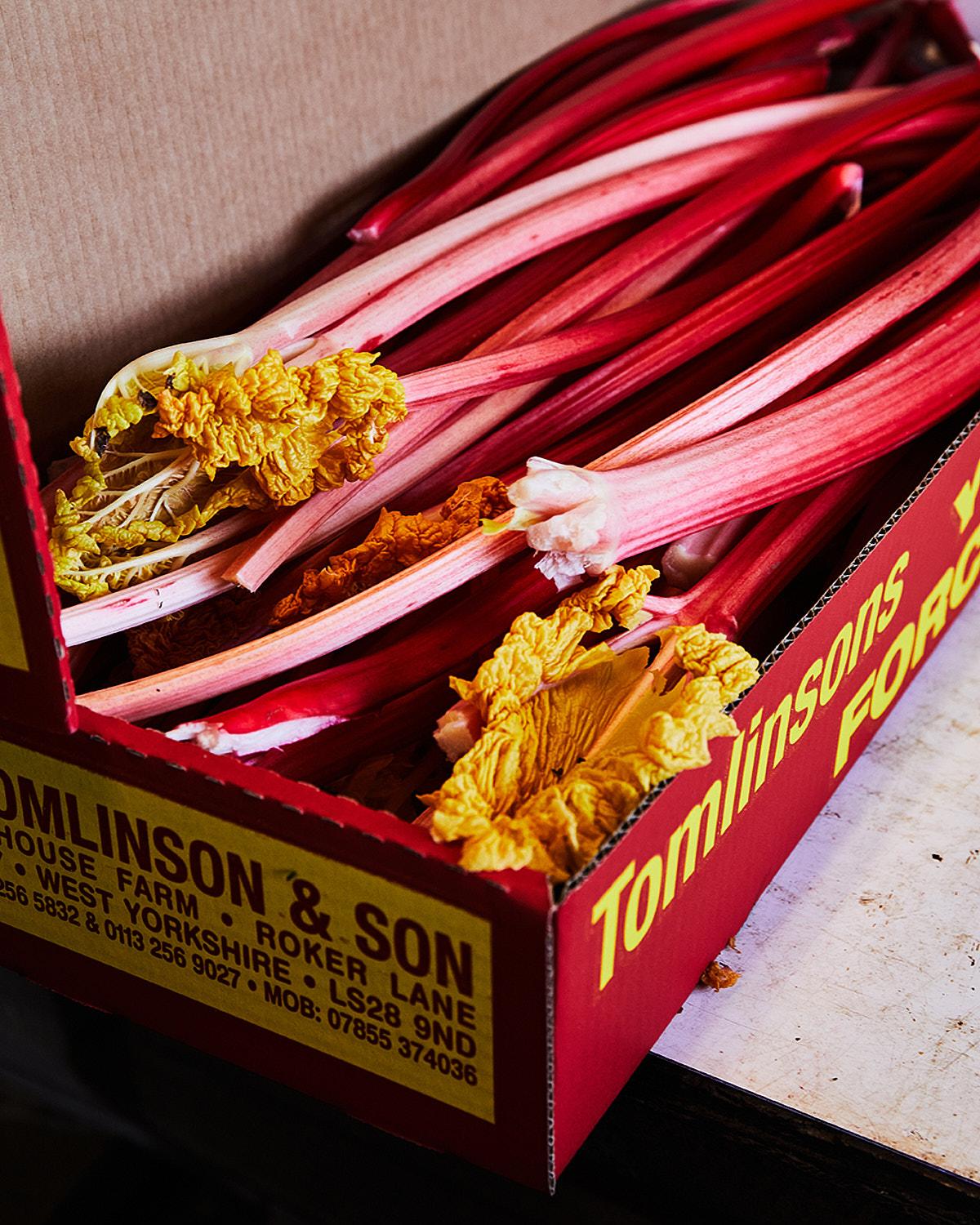 A box containing broken and intact rhubarb stalks with leaves, labeled 'Tomlinson's'.