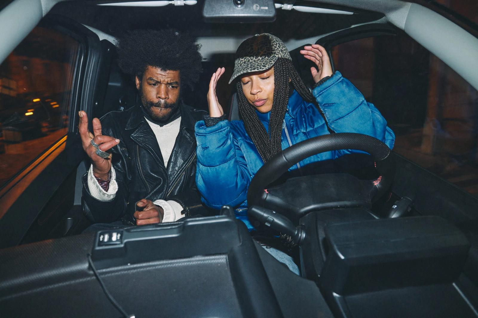 A man and a woman sit in the driver's seat of a car, both looking at the camera with serious expressions. The man is wearing a black leather jacket and has an afro, while the.