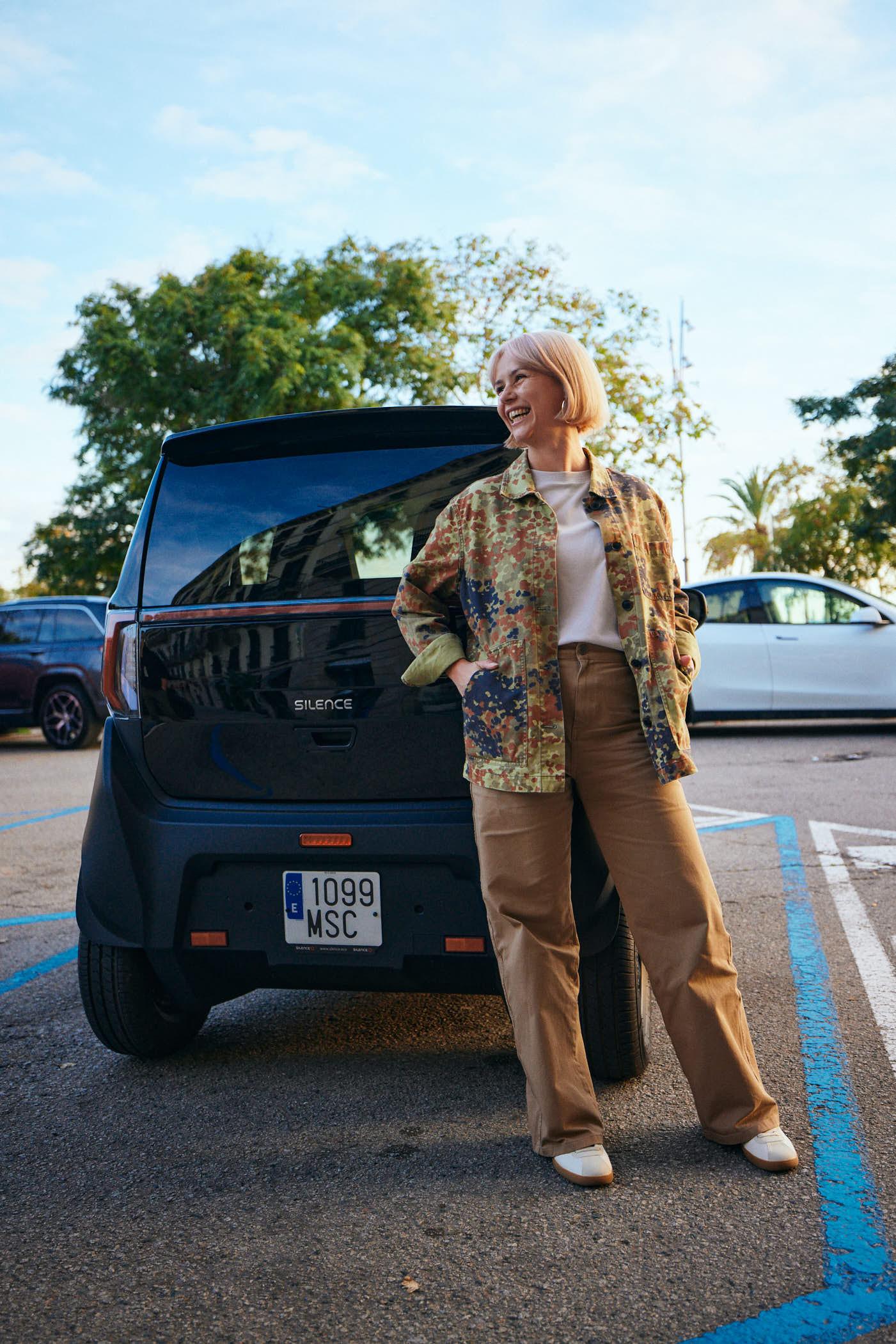 A woman with short blonde hair stands in front of a black car with a white license plate, looking to her left with a smile. She is wearing a colorful, long-sleeved button-down.