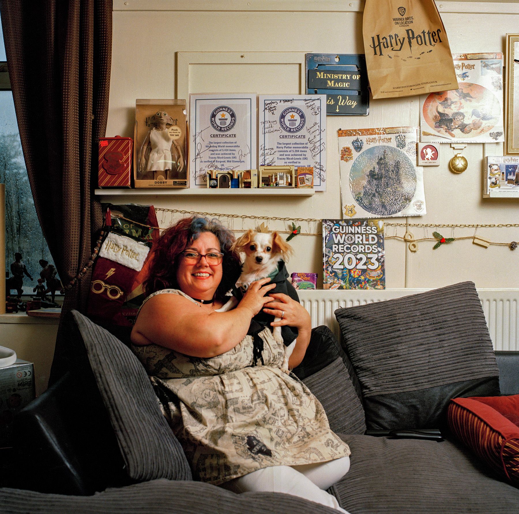 A woman sits on a couch holding a small white dog, with Harry Potter-themed items on the wall behind her.