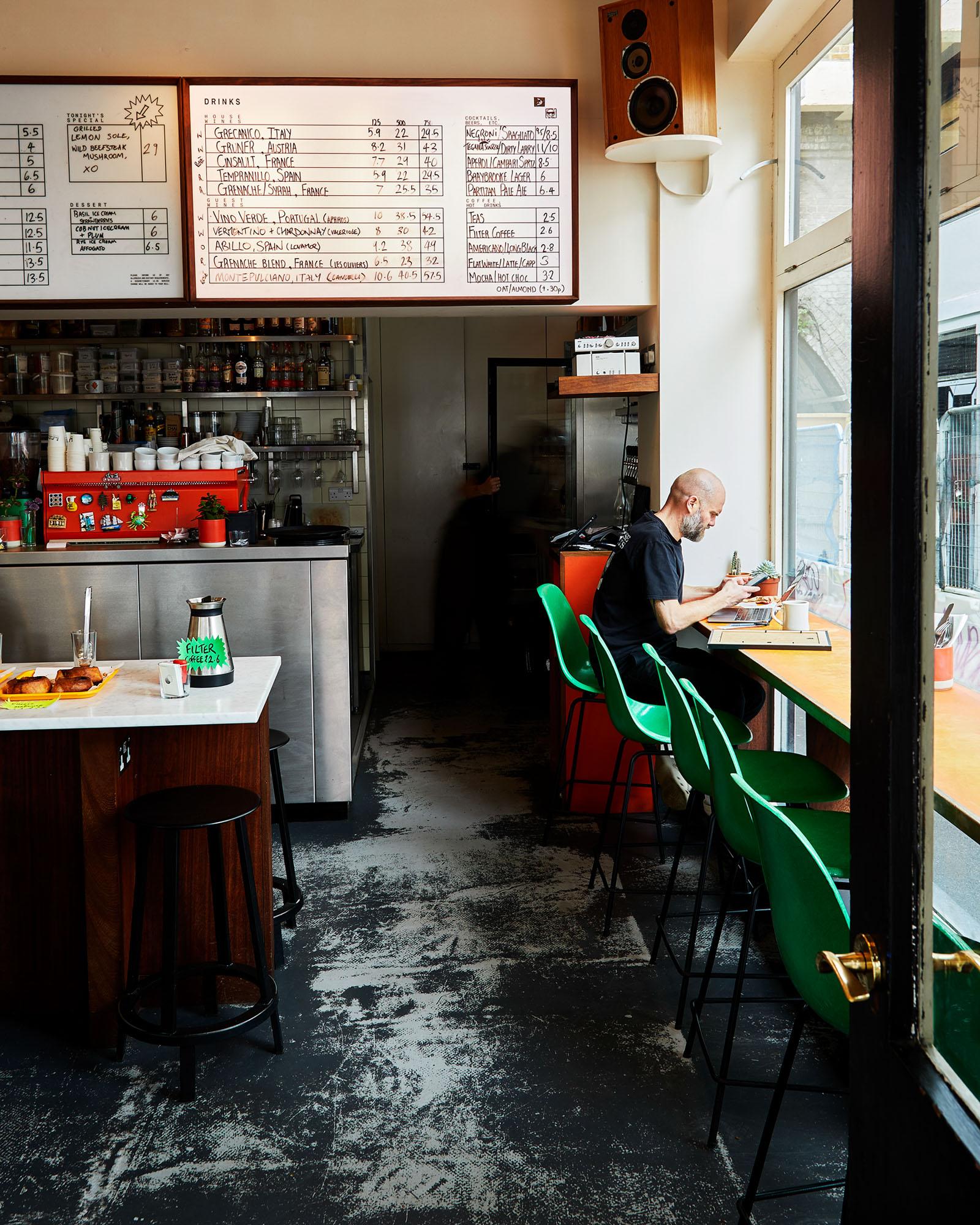 A man sits at a counter in a coffee shop, surrounded by a menu board and a coffee machine.