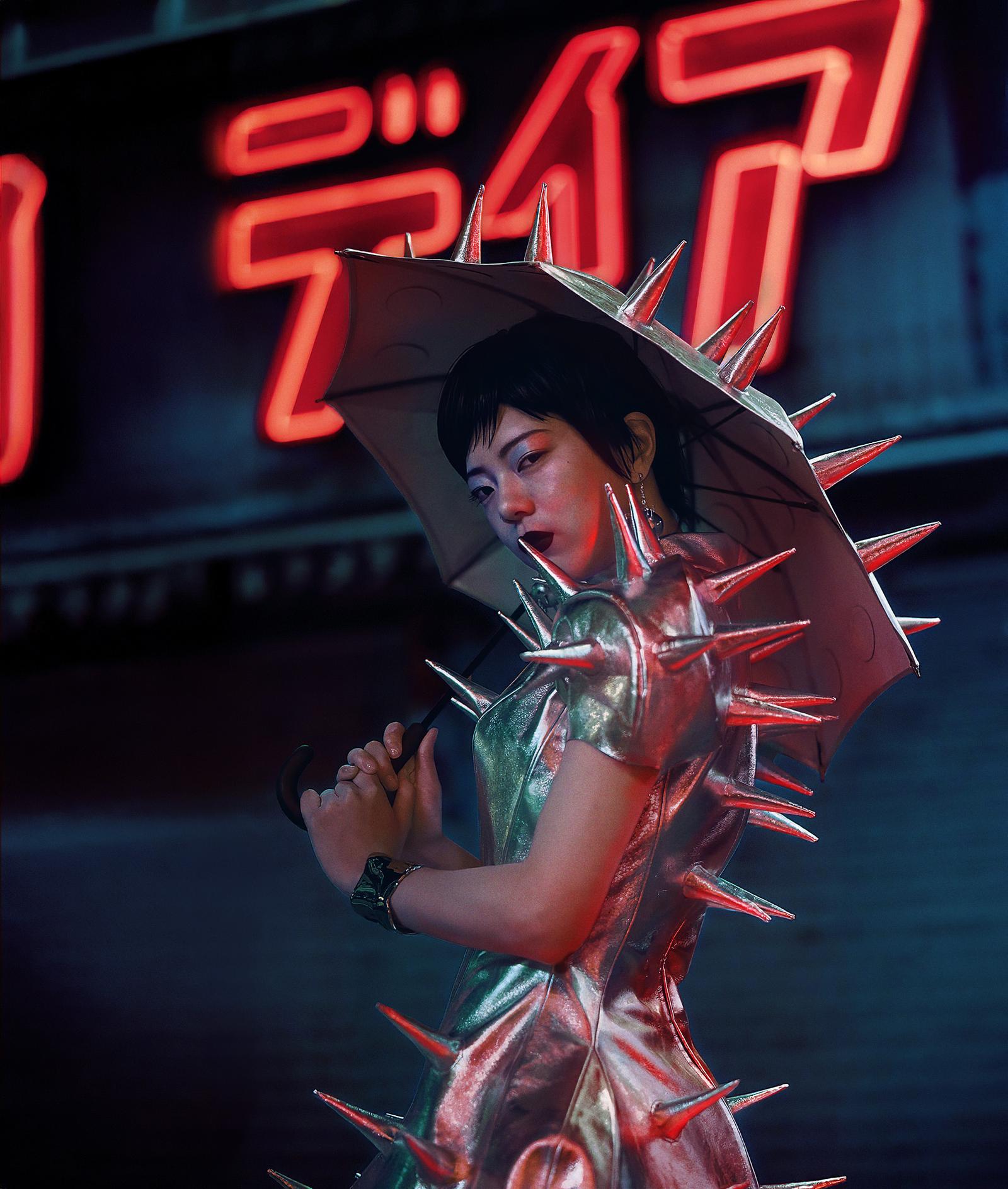 A woman in a futuristic, spiky, metallic dress and umbrella, standing in front of a neon-lit sign in a city at night.