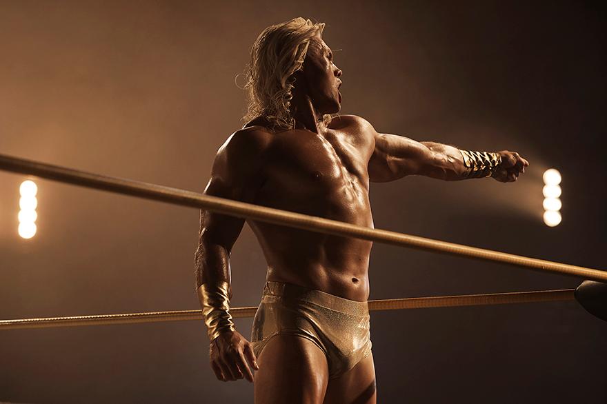 A muscular, blonde, male wrestler in a gold and silver wrestling costume, posing in a wrestling ring.