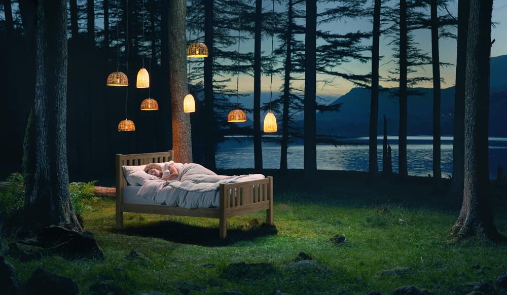 A couple is sleeping peacefully in a bed in a forest clearing overlooking a lake at sunset.