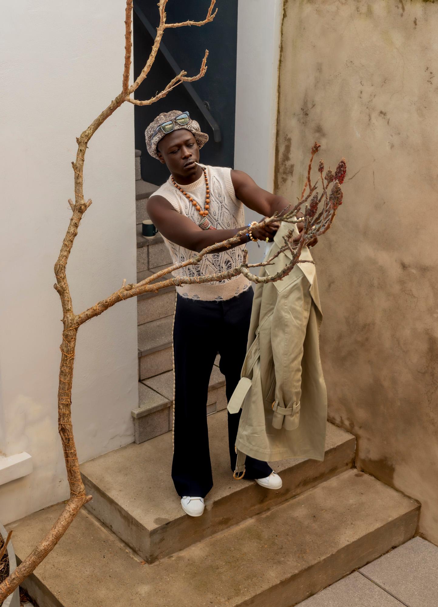 A man in a white tank top, black pants, and a hat, standing on a staircase, holding a beige trench coat.