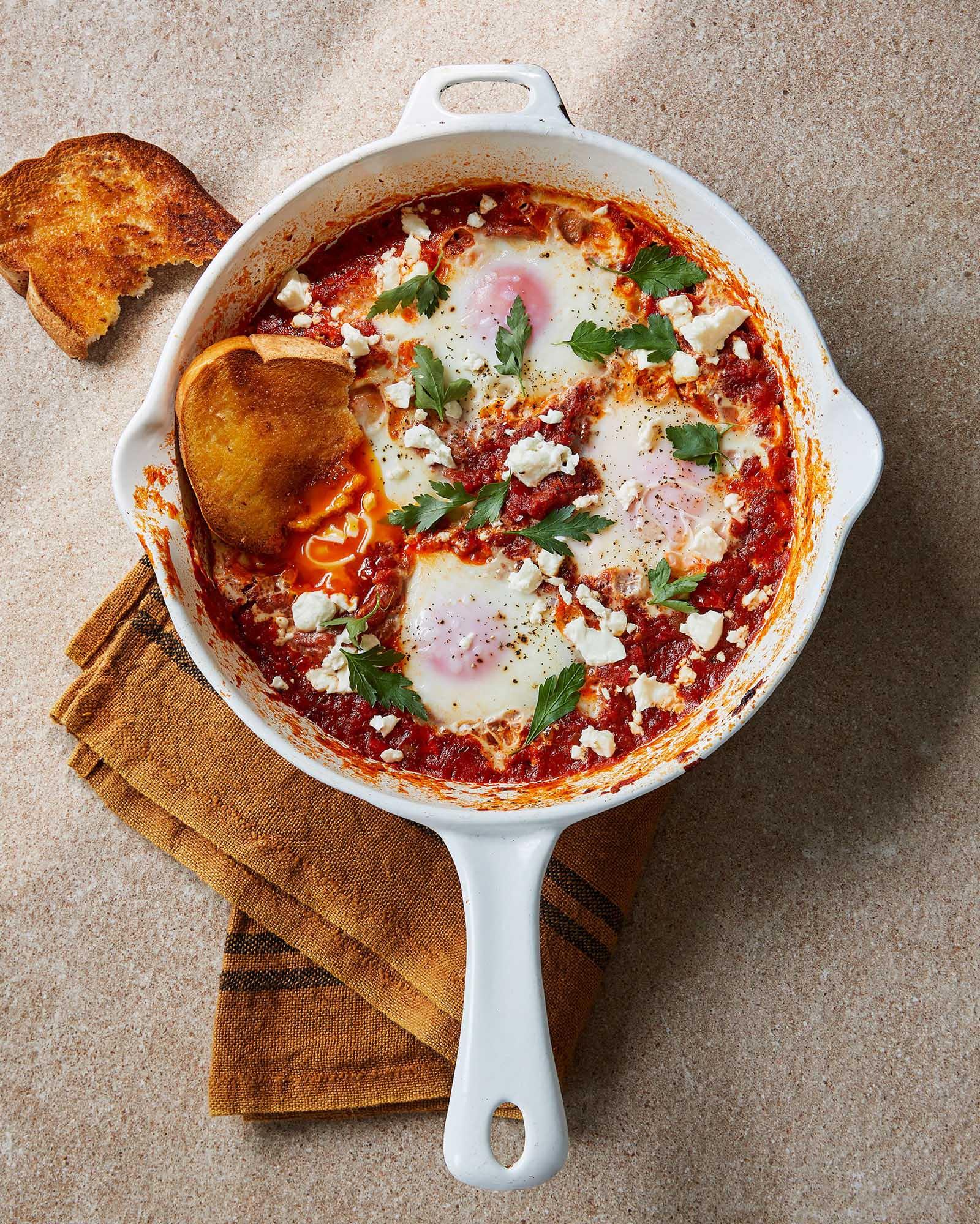 A ceramic skillet with eggs poached in a spicy tomato sauce, topped with feta cheese and parsley, and a slice of toast.