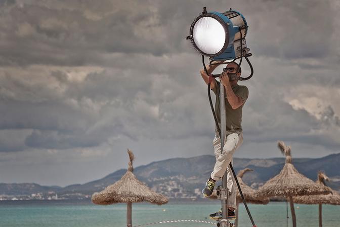 A person in a green t-shirt and white pants is operating a large, blue, industrial-style lighting fixture on a beach with palm trees and a distant city skyline.