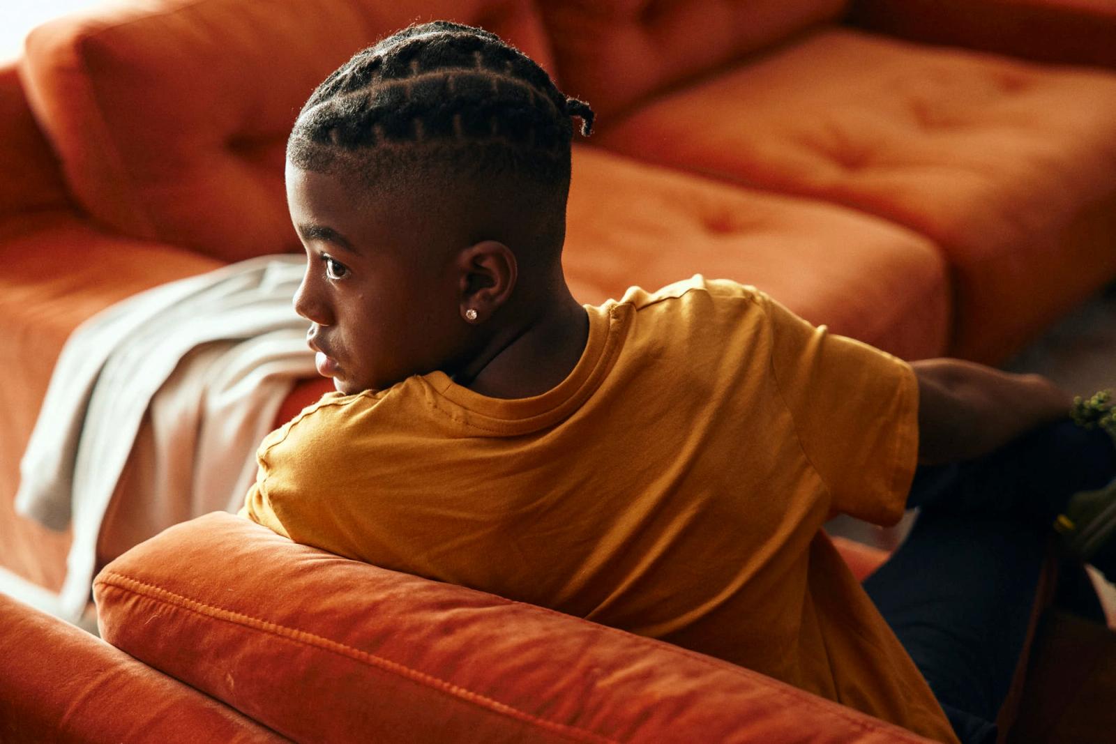 A young black boy with braided hair sits on a couch, looking out of frame to the left. He is wearing a yellow t-shirt and has a serious expression on his face.