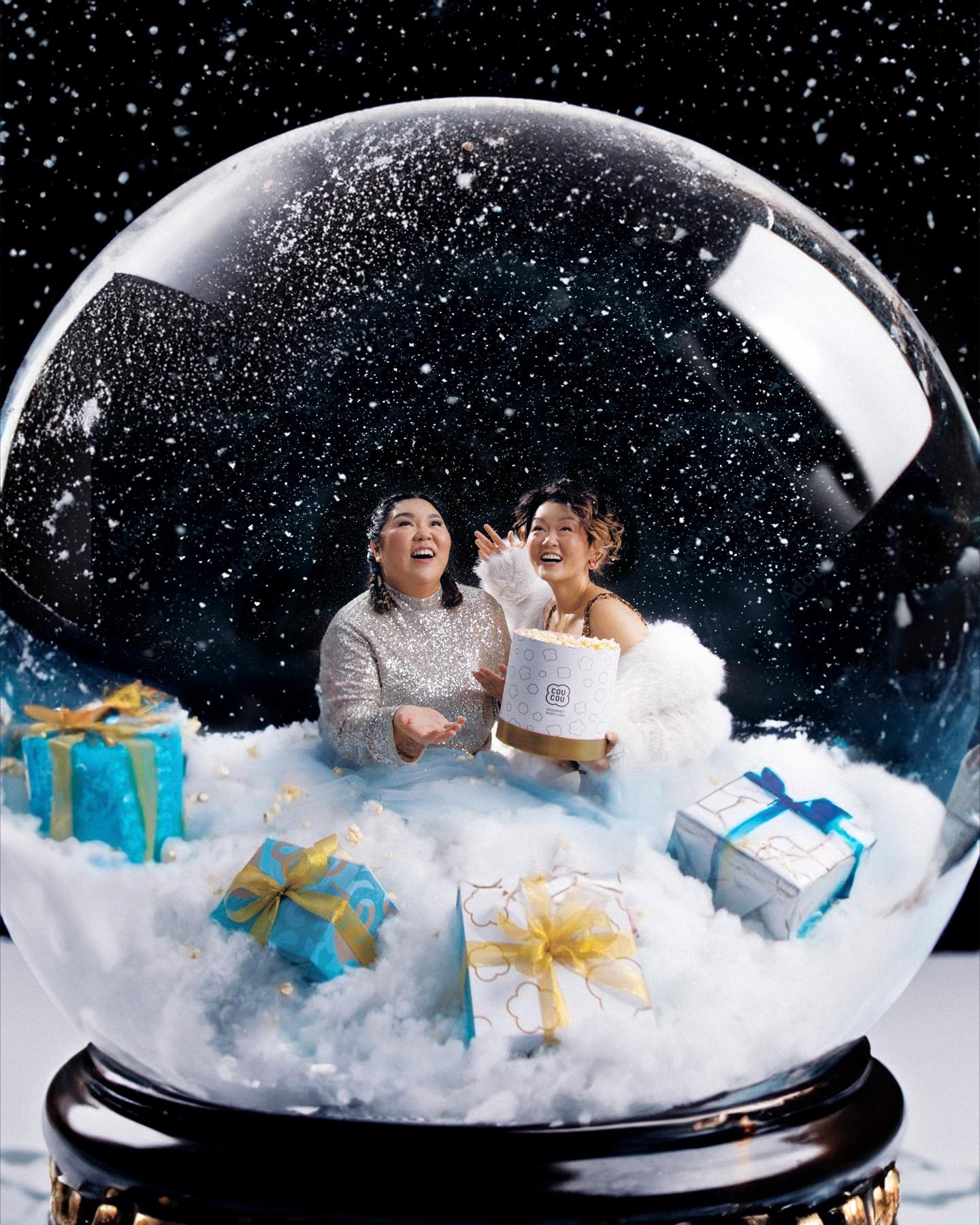 Two women in a snow globe, surrounded by snow and gifts, with a city skyline in the background.